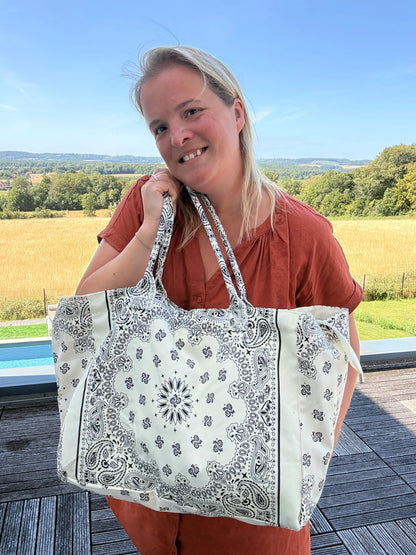 Sophie souriante tenant un grand sac polochon blanc et noir à motif bandana paisley, en extérieur près d’un paysage champêtre.