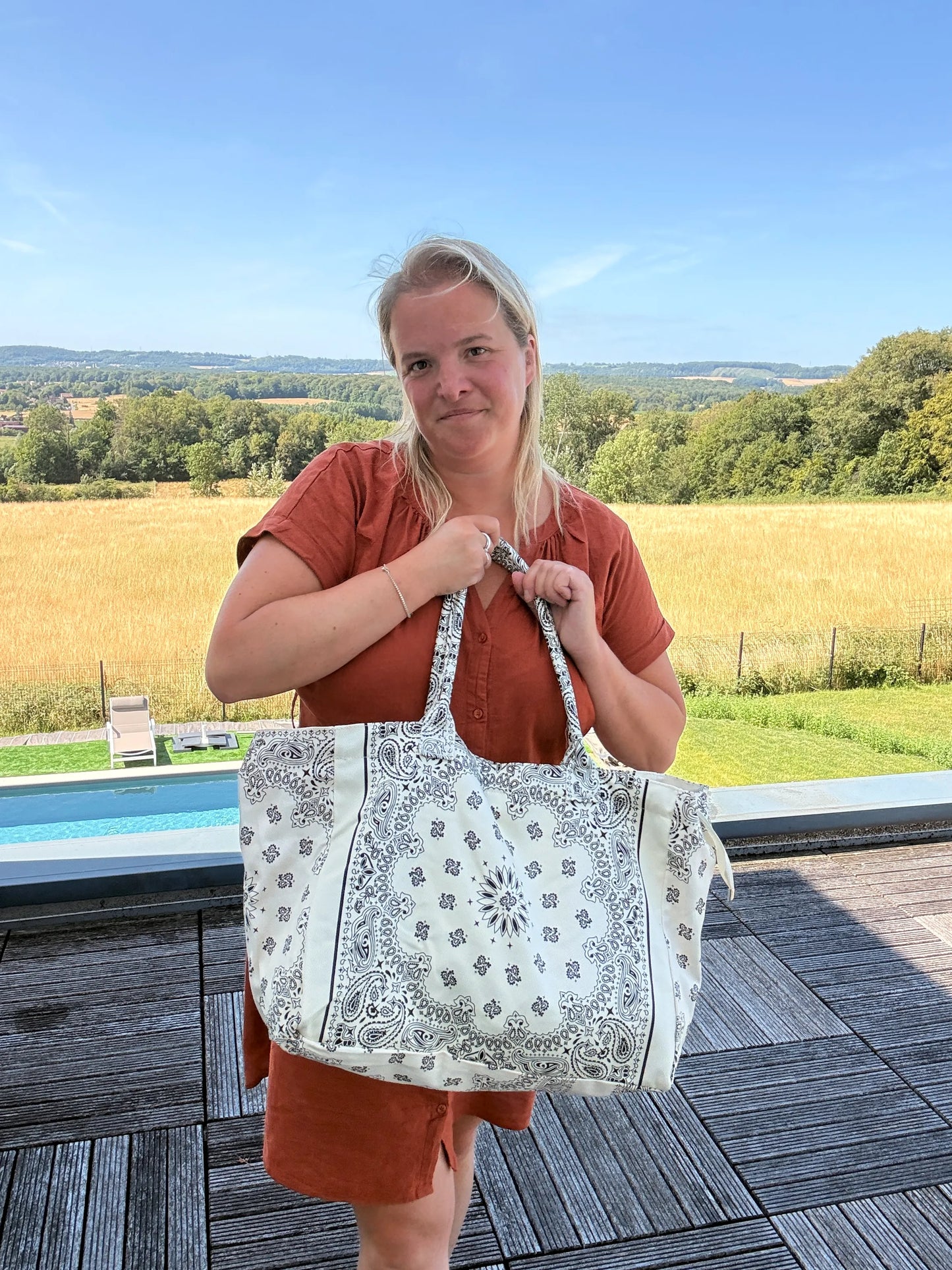 Homme ou femme portant un grand sac polochon blanc à motifs paisley, debout en extérieur près d’une piscine, paysage champêtre en arrière-plan. Grand Sac Polochon Paisley Blanc – Le Sac Bohème Chic de Toutes les Saisons.