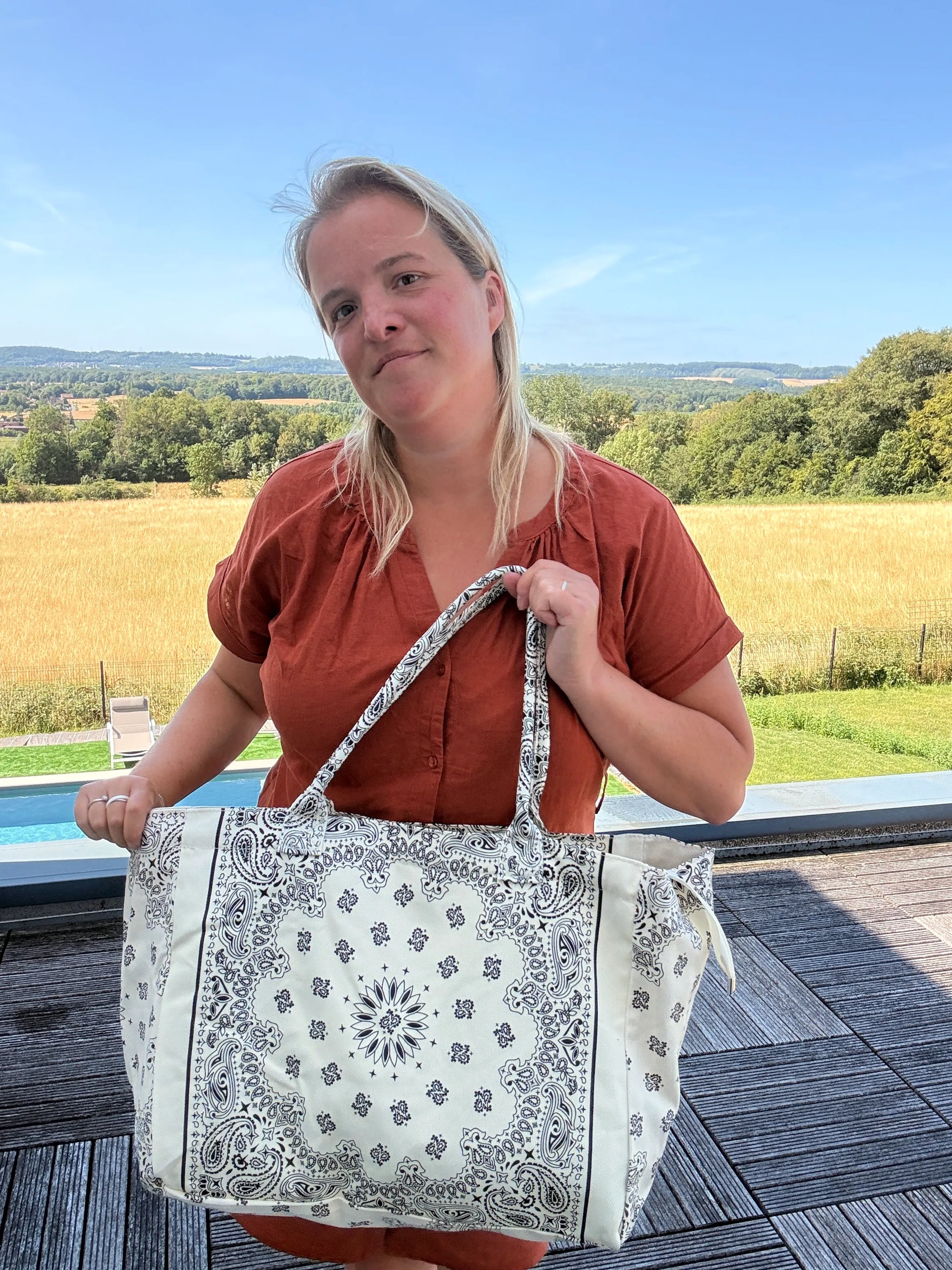 Portrait d’une femme tenant un grand sac polochon blanc à motif paisley, en extérieur sur une terrasse avec champ et ciel bleu. Grand sac bohème chic.