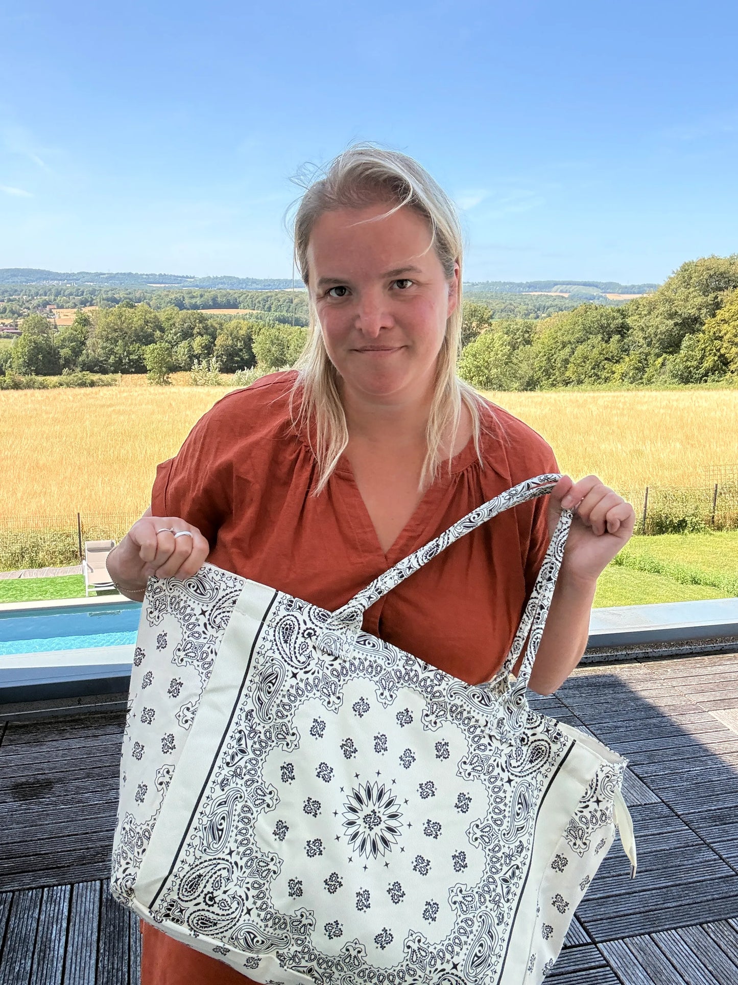 Grande sac polochon paisley blanc porté par une jeune femme en robe brique, fond champ et villa, motif bandana noir sur fond blanc. Grand sac bohème chic toutes saisons.