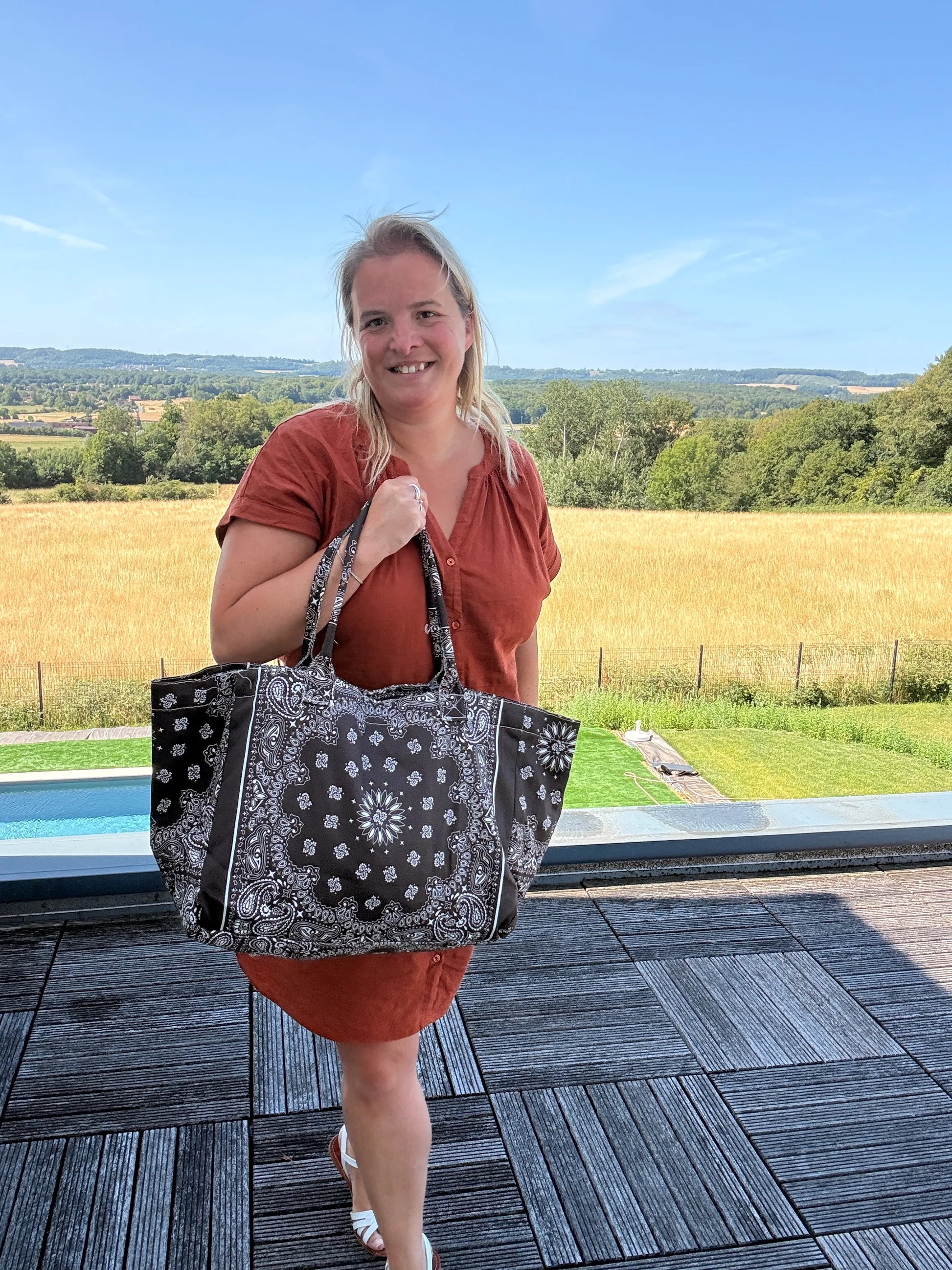 Homme ou femme debout sur une terrasse, portant un grand sac polochon marron à motif bandana noir et blanc, paysage rural en arrière-plan, ciel bleu.