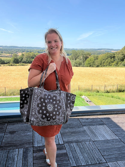 Homme ou femme debout sur une terrasse, portant un grand sac polochon marron à motif bandana noir et blanc, paysage rural en arrière-plan, ciel bleu.
