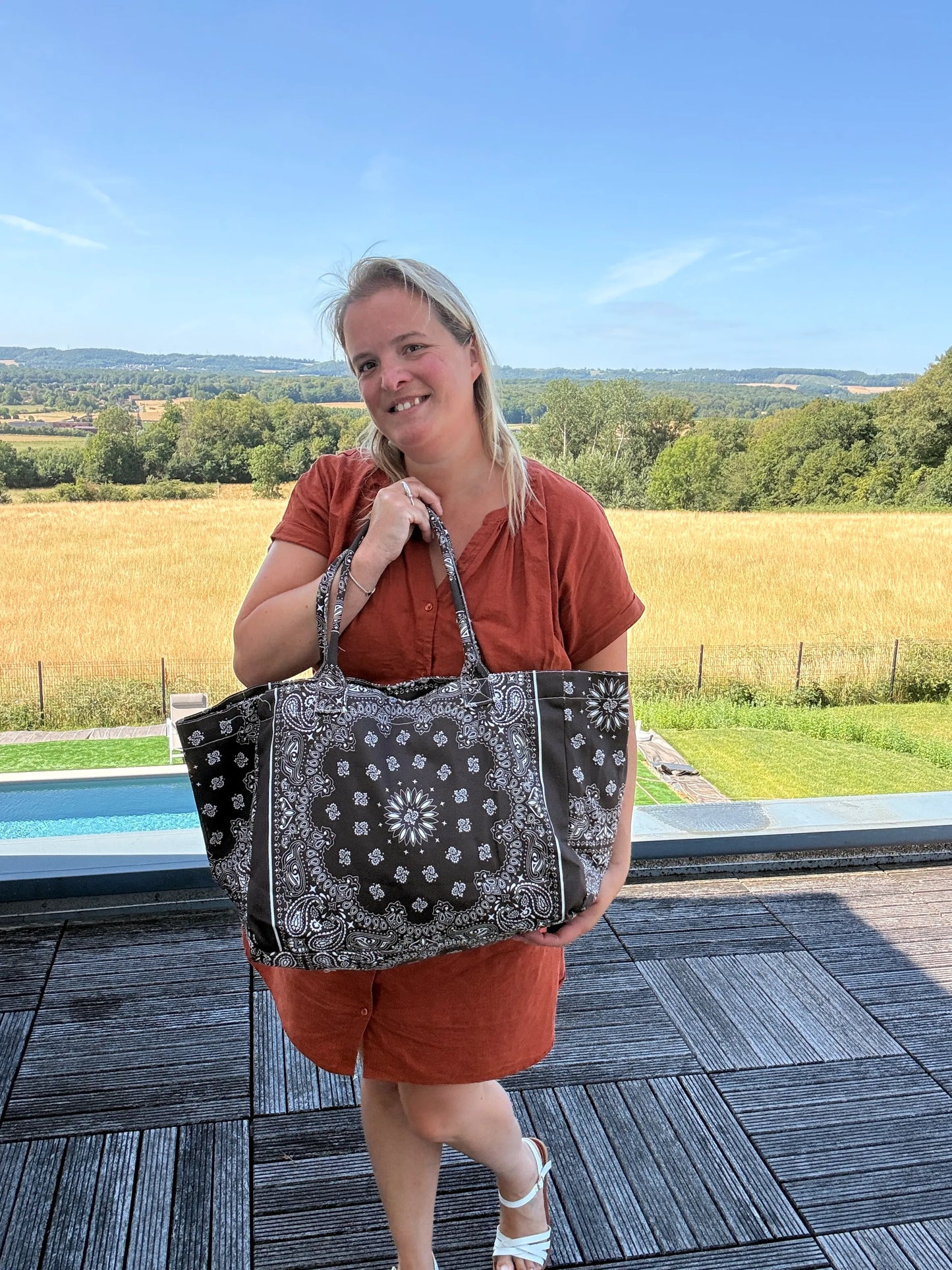 Femme debout sur une terrasse rurale, portant un sac polochon noir à motifs bandana, manches courtes orange, paysage champêtre en arrière-plan.