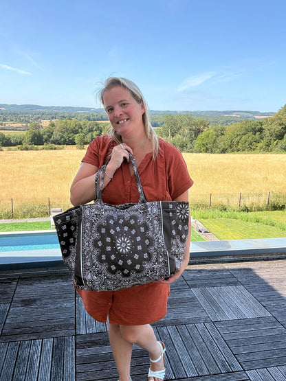 Femme debout sur une terrasse rurale, portant un sac polochon noir à motifs bandana, manches courtes orange, paysage champêtre en arrière-plan.