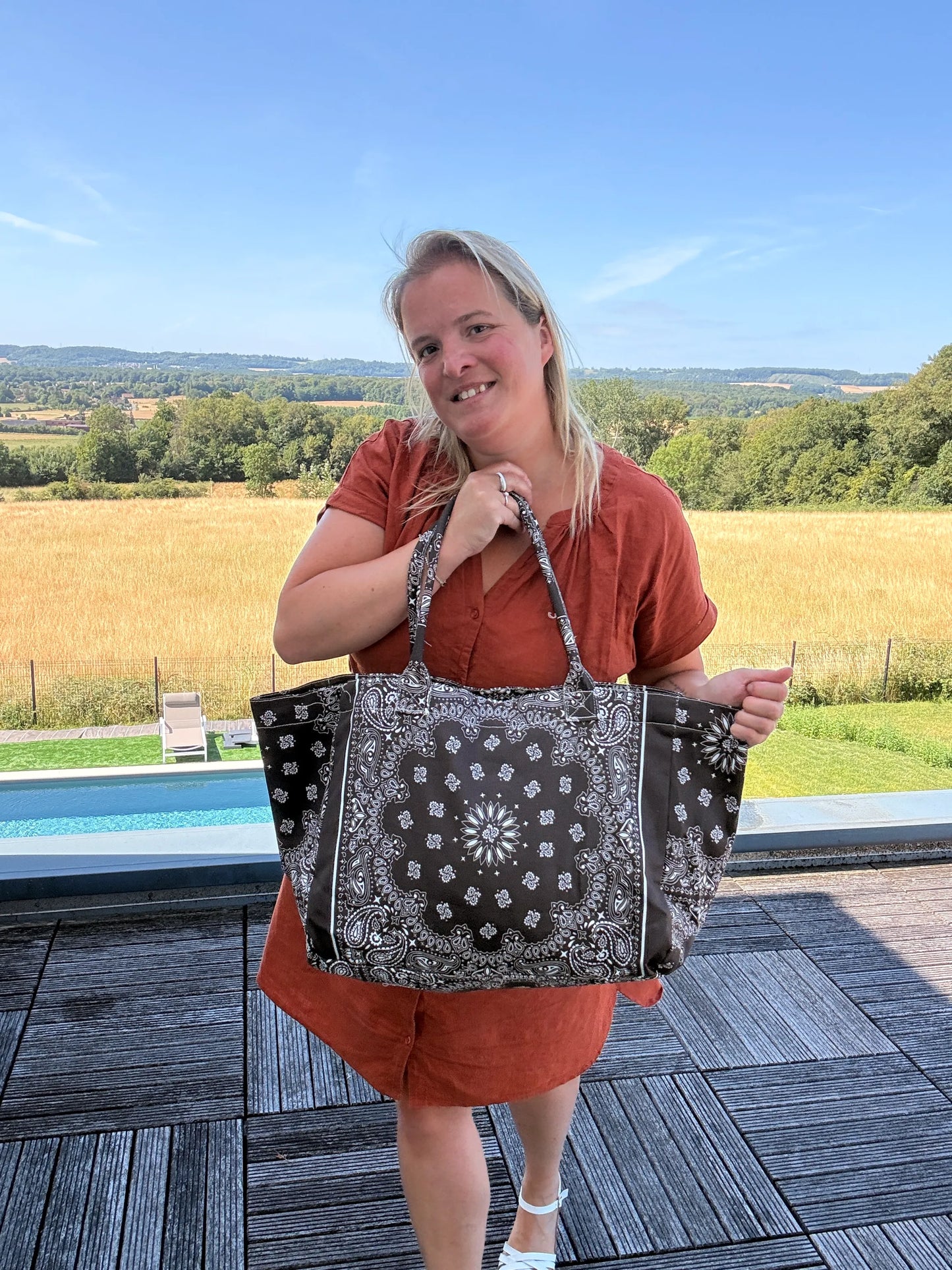 Grand sac polochon marron zippé porté par une femme sur une terrasse extérieure, paysage champêtre en arrière-plan.