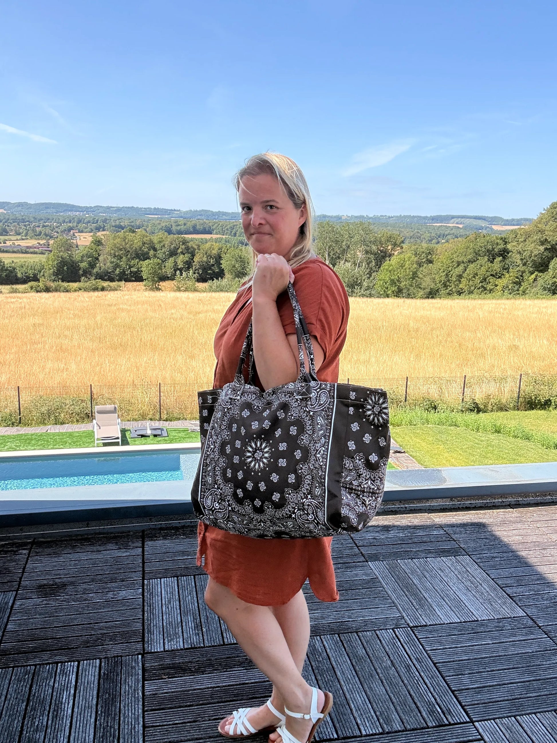 Grand sac polochon marron zippé porté par une femme près d’une piscine, campagne en arrière-plan, look chic et décontracté.