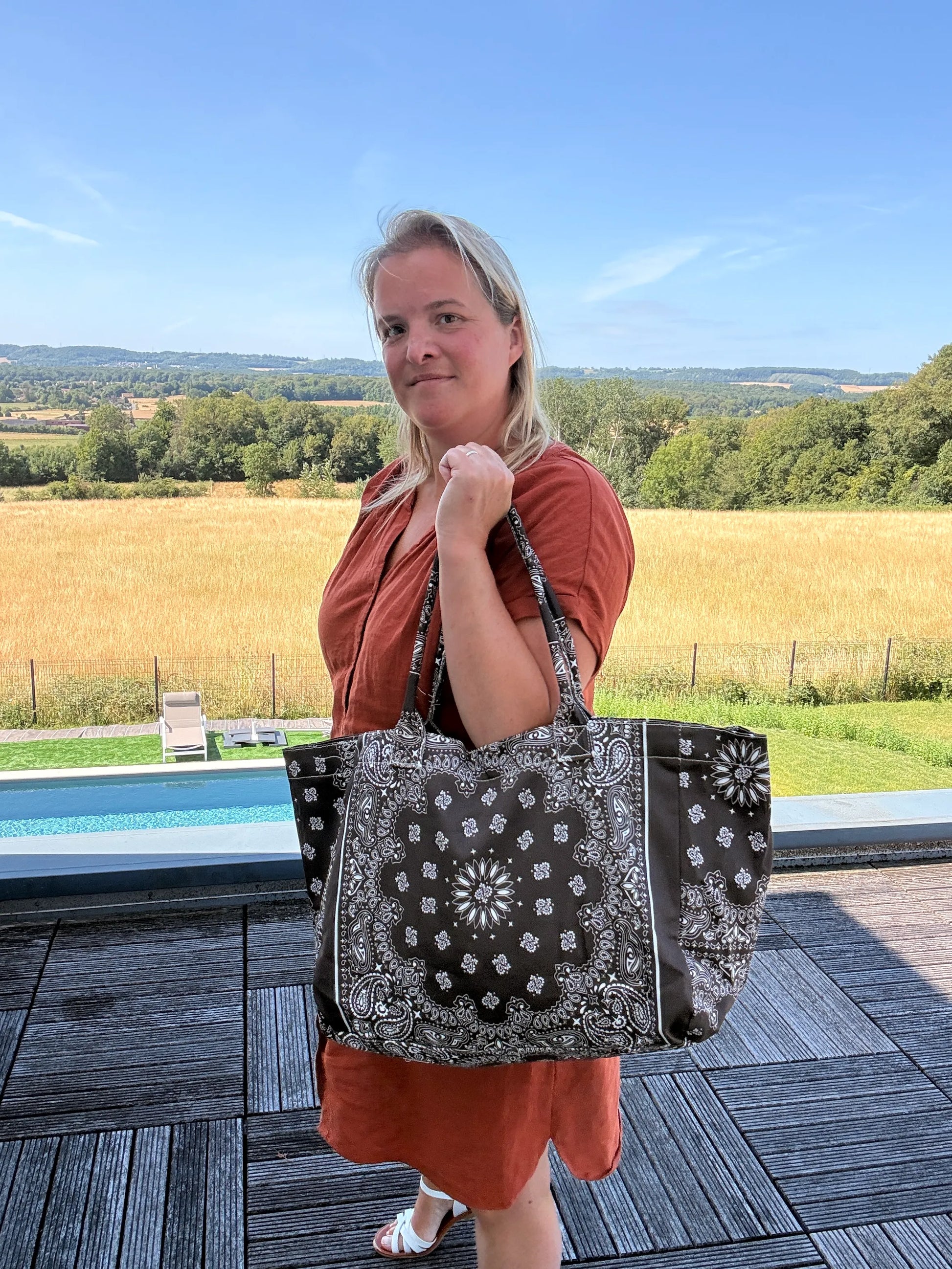 Homme ou femme portant un grand sac polochon noir à motif bandana blanc, sur une terrasse près d’une piscine, champ en arrière-plan, en robe orange et sandales.