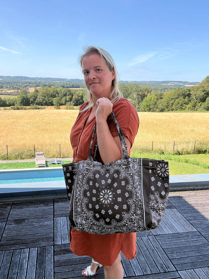 Homme ou femme portant un grand sac polochon noir à motif bandana blanc, sur une terrasse près d’une piscine, champ en arrière-plan, en robe orange et sandales.