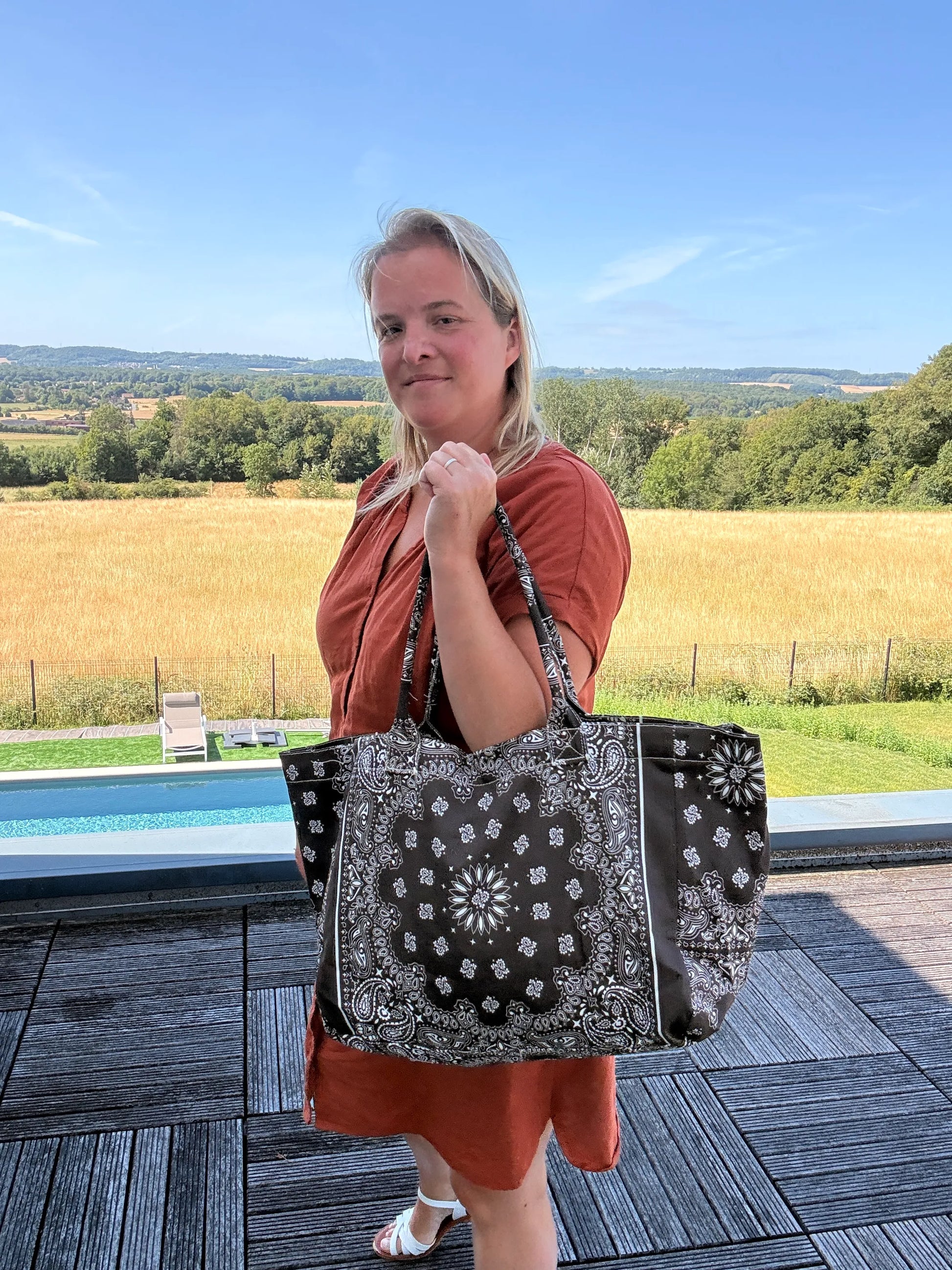 Grand sac polochon marron zippé porté par une femme près d’une piscine, paysage rural en arrière-plan.