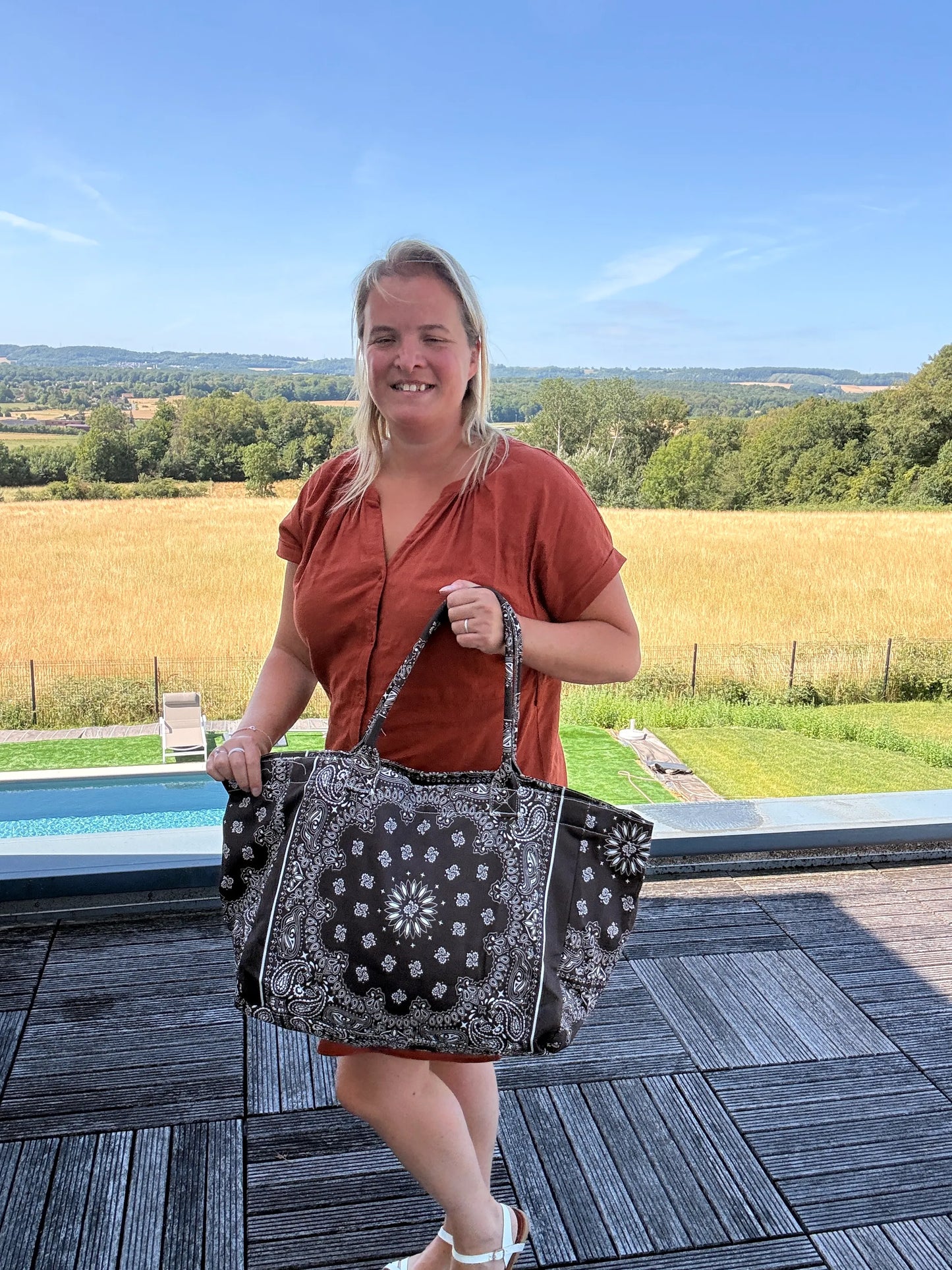 Grande sac polochon marron zippé porté par une femme en terrasse en plein air, paysage champêtre en arrière-plan.