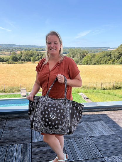 Grande sac polochon marron zippé porté par une femme en terrasse en plein air, paysage champêtre en arrière-plan.