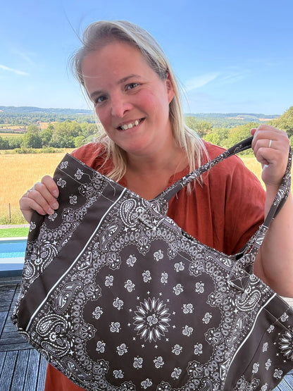 Femme souriante tenant un grand sac polochon marron à motifs bandana noir et blanc en extérieur.