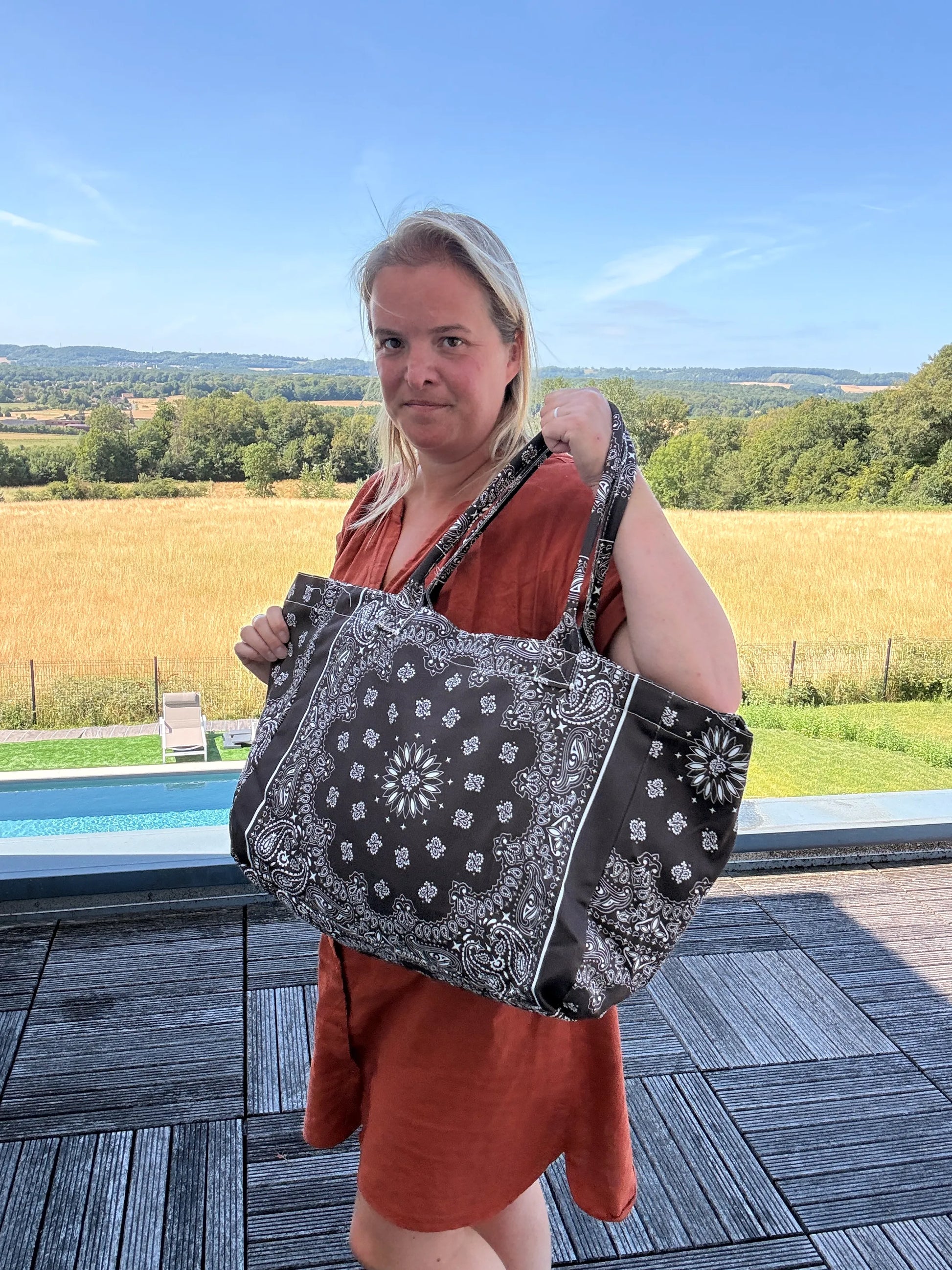 Grande sac polochon noir à motifs bandana porté par une femme en robe orange, devant piscine et paysage champêtre.