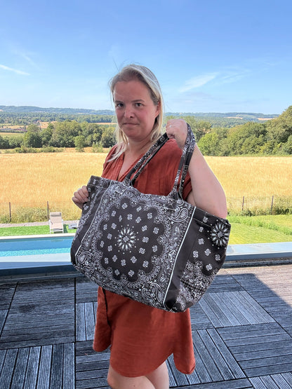 Grande sac polochon noir à motifs bandana porté par une femme en robe orange, devant piscine et paysage champêtre.