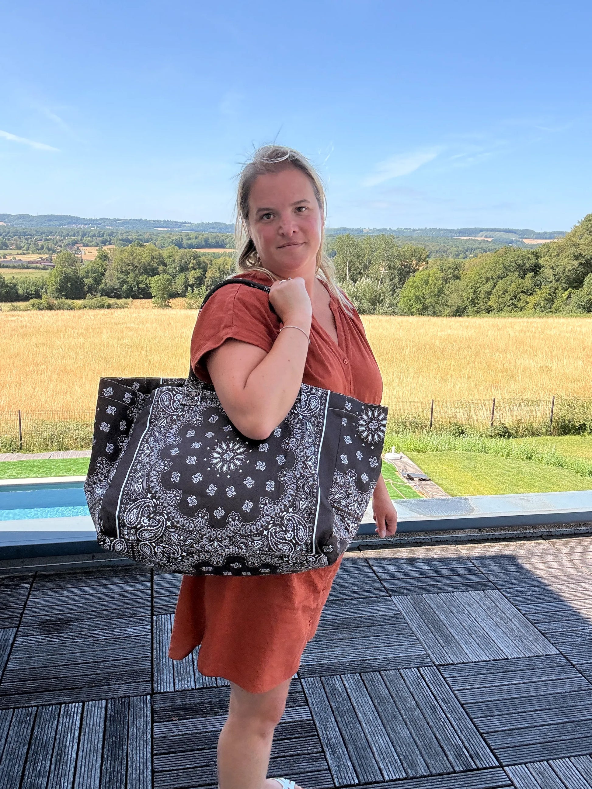 Homme ou femme portant un grand sac polochon noir à motifs paisley, sur une terrasse extérieure avec champ et ciel bleu.