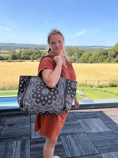 Homme ou femme portant un grand sac polochon noir à motifs paisley, sur une terrasse extérieure avec champ et ciel bleu.