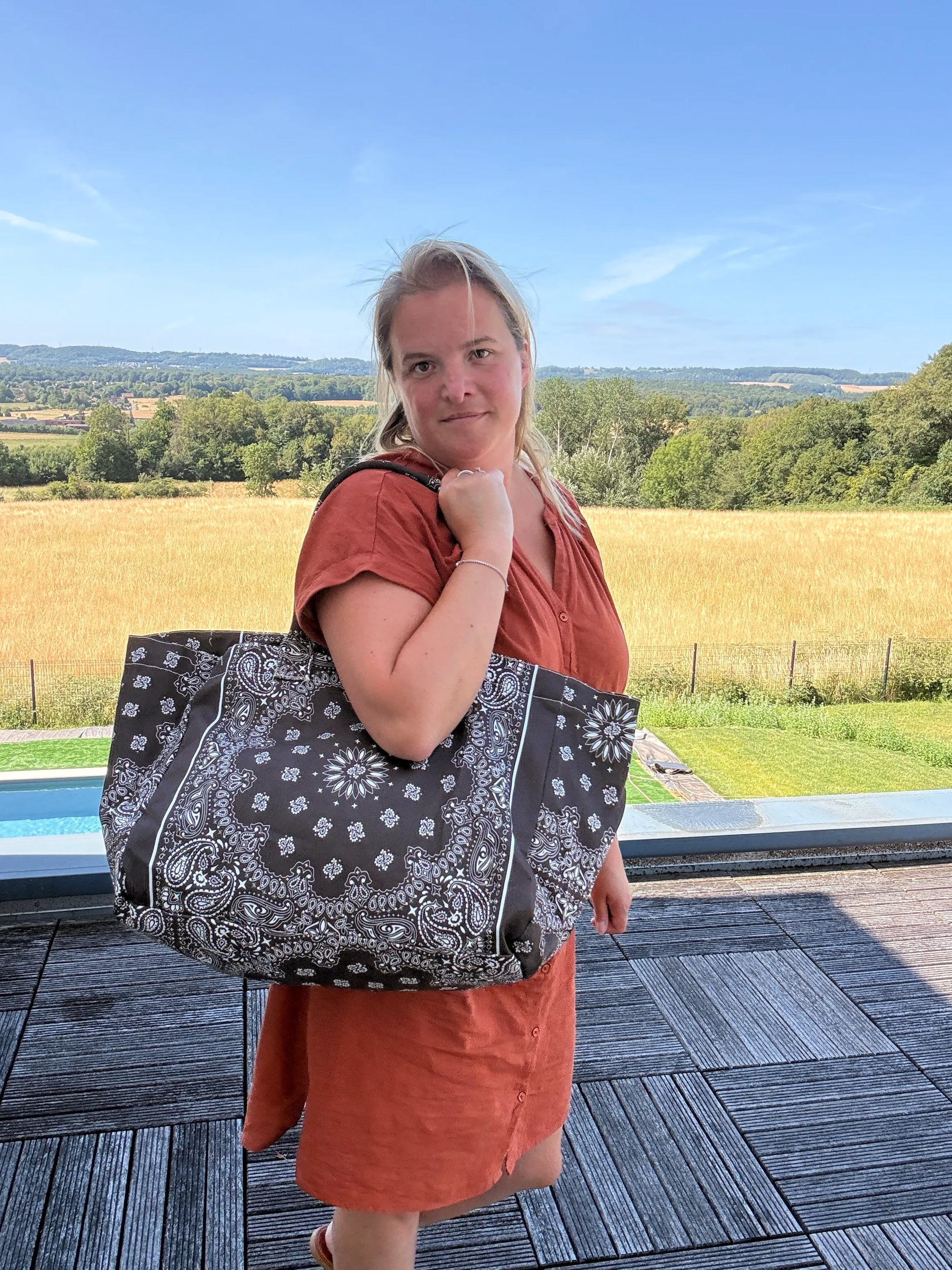 Grand sac polochon marron zippé porté sur l’épaule d’une femme en robe orange, décor champêtre en arrière-plan et paysage verdoyant.