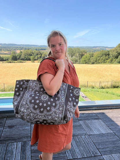 Grand sac polochon marron zippé porté sur l’épaule d’une femme en robe orange, décor champêtre en arrière-plan et paysage verdoyant.