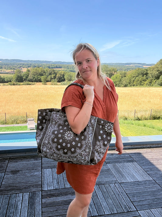 Grande femme portant un sac polochon marron zippé à motif bandana noir près d’une piscine et d’un paysage champêtre.