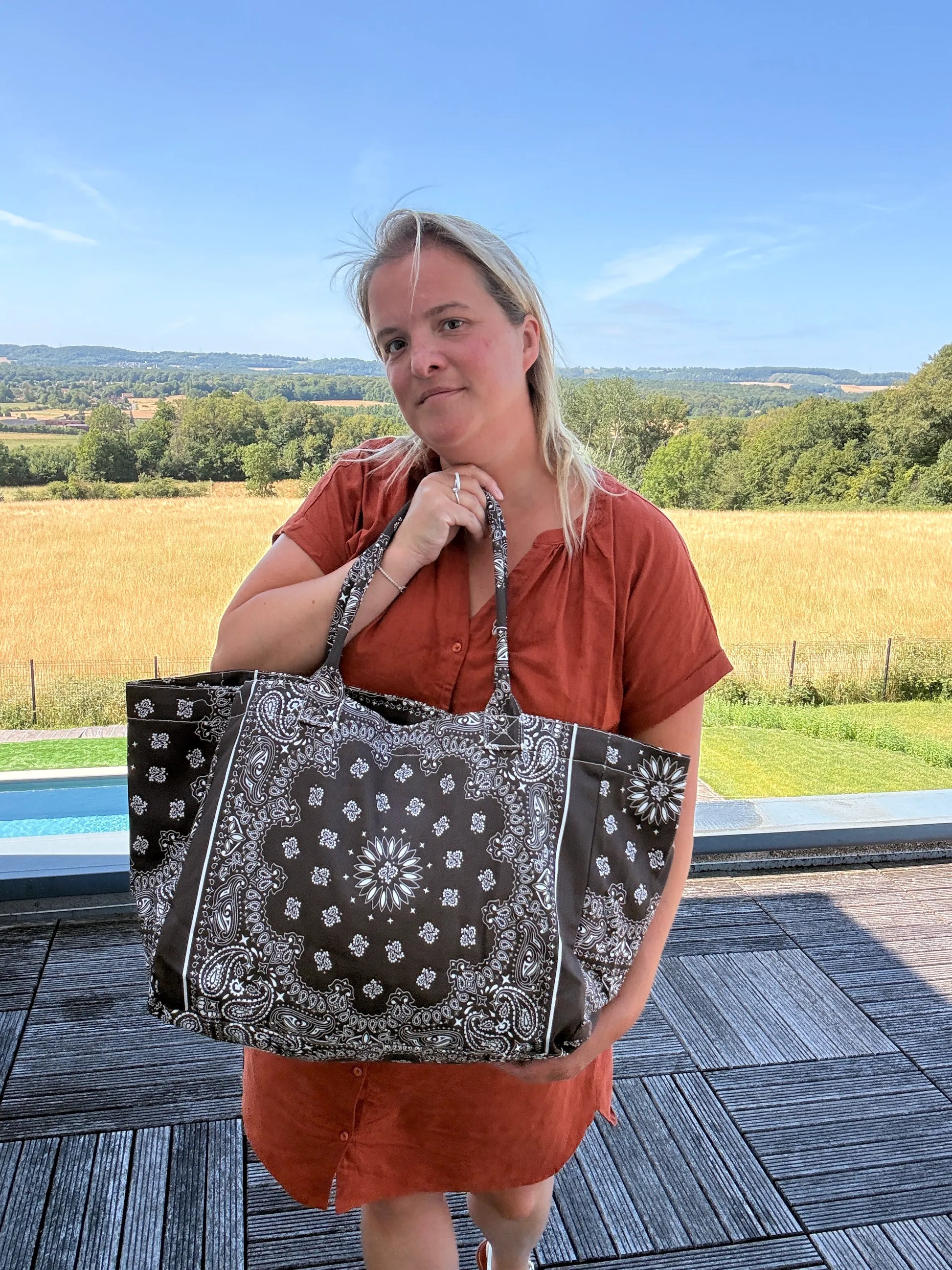 Grand sac polochon marron zippé porté par une femme en robe orange, en extérieur, montrant son grand format et sa bandoulière.