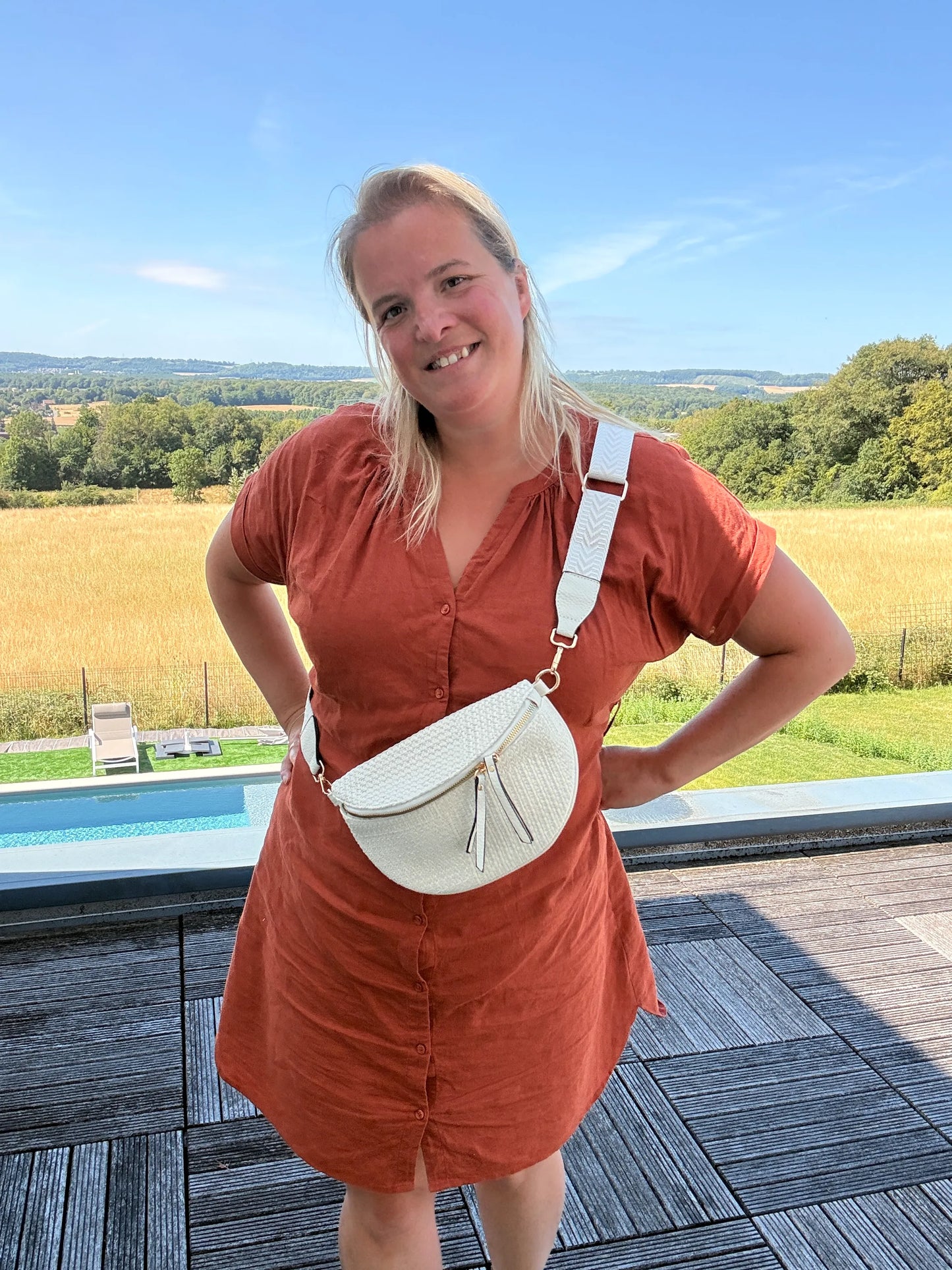 Sac banane bohème pour femme portée en bandoulière, couleur beige écru avec raphia, sur fond rural en été.