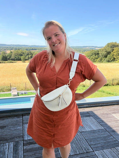 Sac banane bohème pour femme portée en bandoulière, couleur beige écru avec raphia, sur fond rural en été.