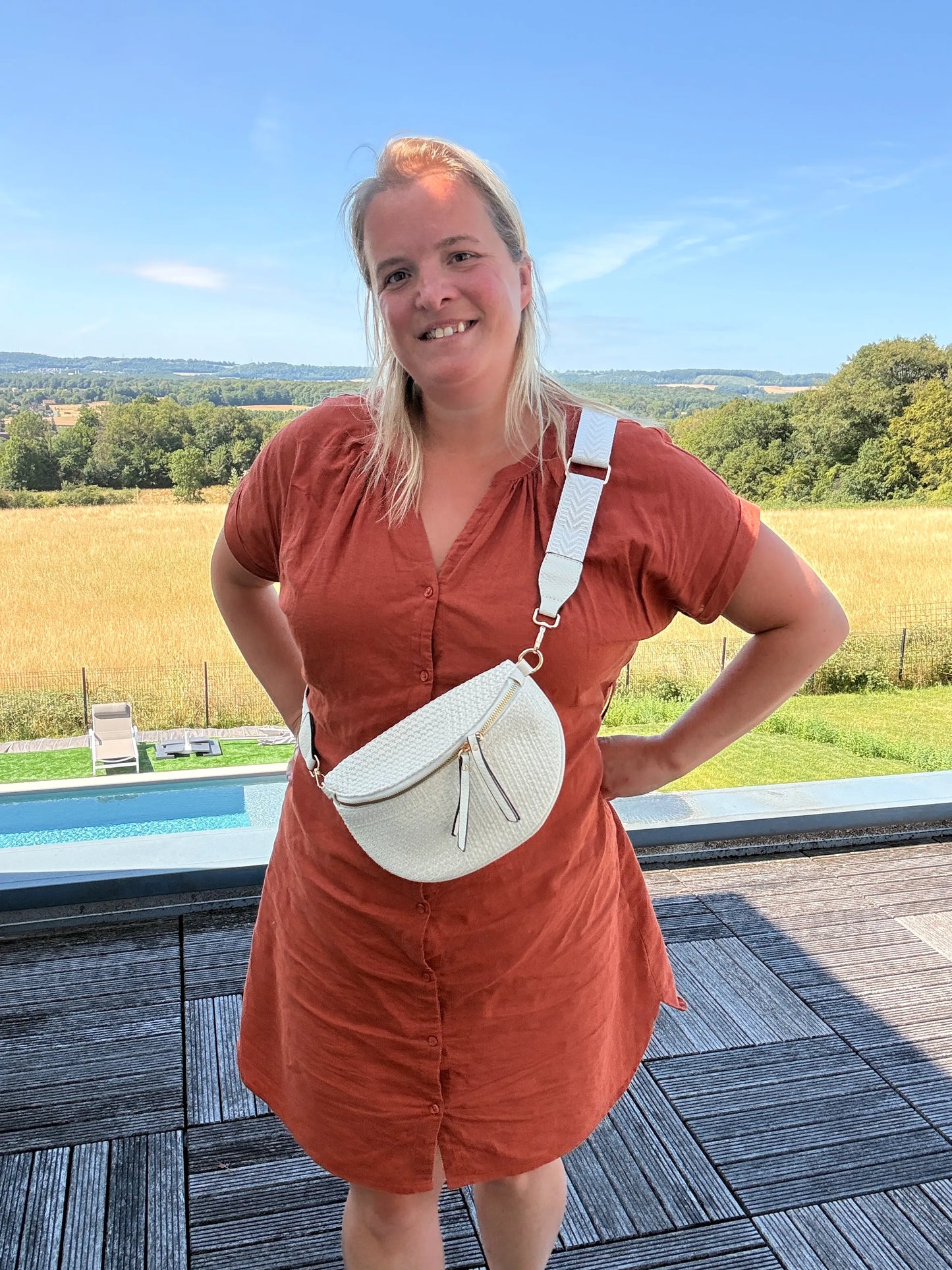 Homme? Non: femme portant sac banane bohème beige en paille raphia, bandoulière réglable, debout en extérieur avec paysage champêtre.