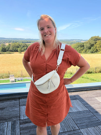 Homme? Non: femme portant sac banane bohème beige en paille raphia, bandoulière réglable, debout en extérieur avec paysage champêtre.