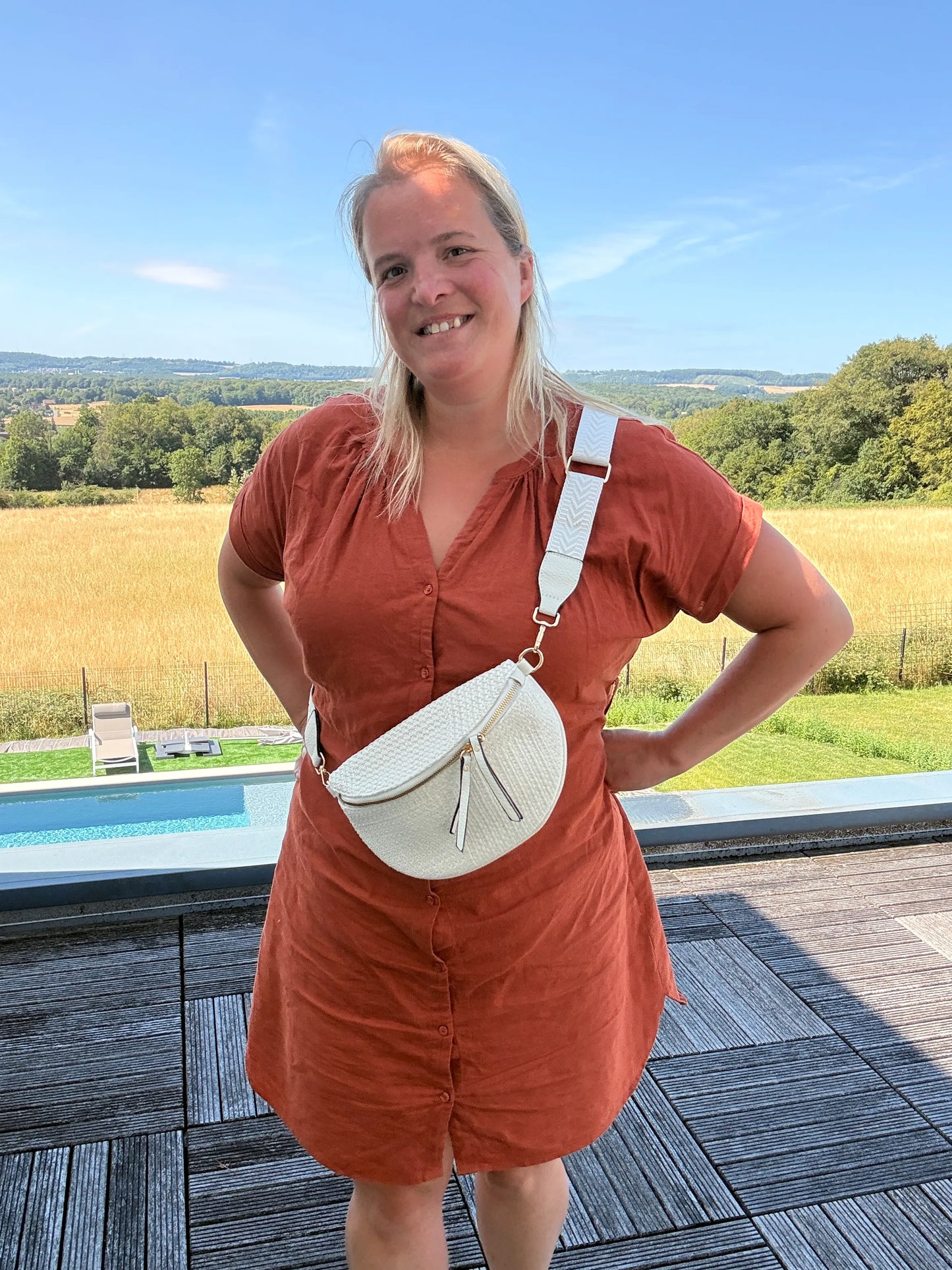 Sac banane bohème femme en paille raphia beige, portée latchée croisée sur l’épaule avec bandoulière réglable, devant paysage champêtre.