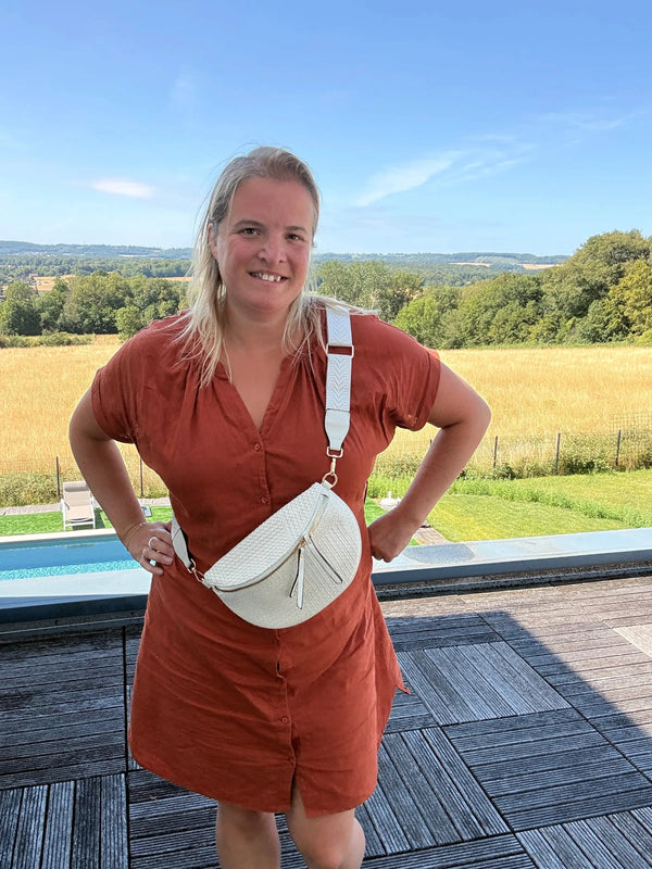 Sac banane bohème pour femme en paille et raphia beige, portée en bandoulière réglable sur une robe orange devant paysage champêtre.