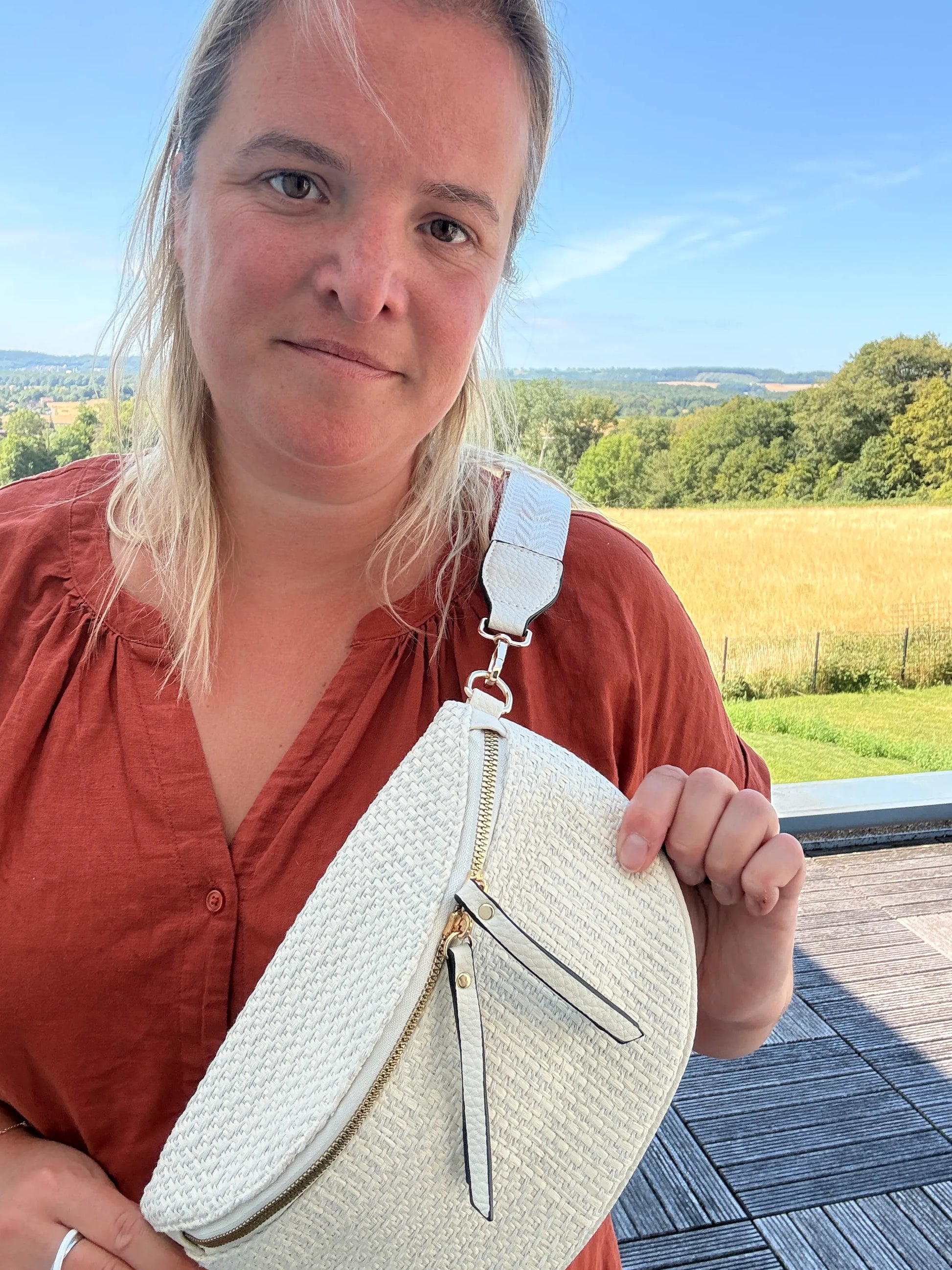 GIRL en chemise rouge tient un sac banane Bohème Femme en paille raphia beige avec bandoulière réglable blanche, fond paysage champêtre.