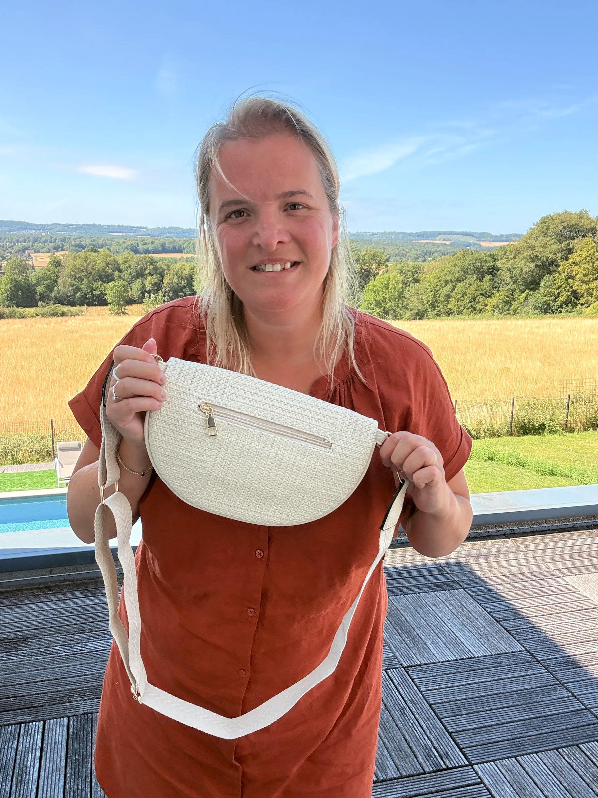 Homme ou femme tenant un sac banane Bohème femme – Paille raphia beige or bandoulière réglable, avec arrière-plan champ et piscine.