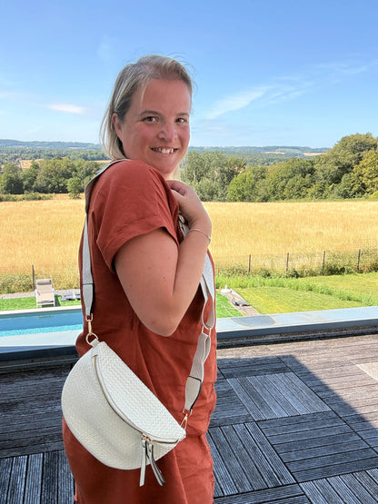 Sac banane bohème femme en paille raphia beige avec bandoulière réglable porté en hauteur par une femme en bordeaux, paysage champêtre en arrière-plan.