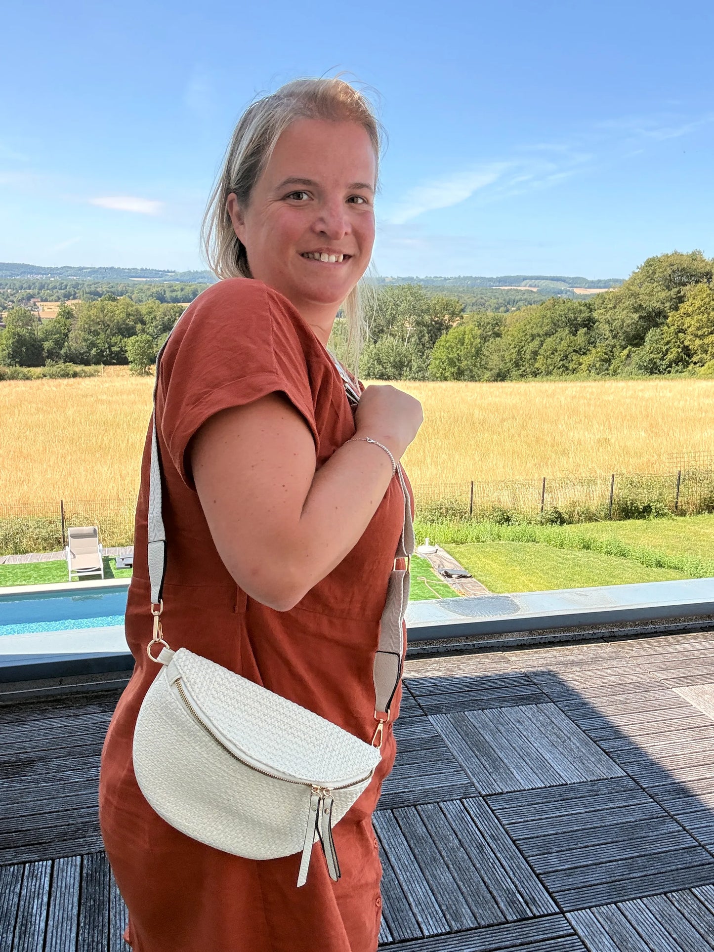 Sac Banane Bohème Femme – femme portant sac banane beige en paille raphia avec bandoulière réglable sur fond extérieur en prairie et piscine.
