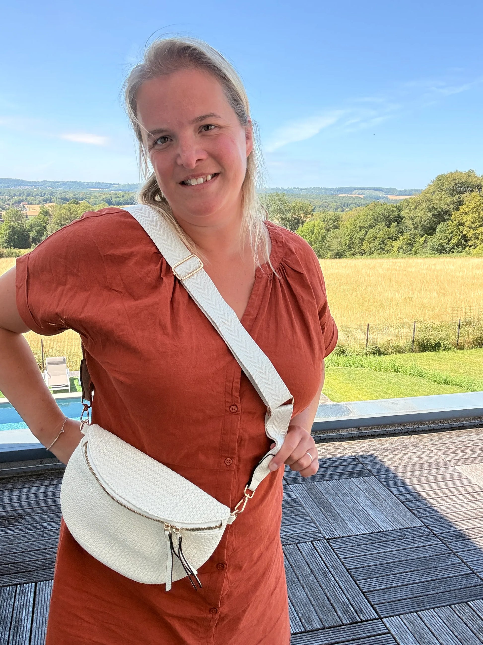 Sac banane bohème femme en paille raphia beige avec bandoulière réglable portée en diagonale sur l’épaule, près d’un terrain champêtre.