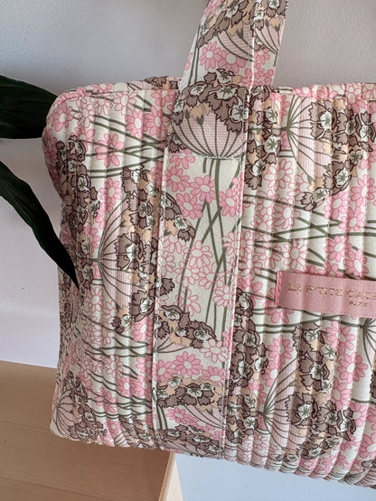Sac polochon matelassé rose fleuri, bandoulière et texte discret sur étiquette, motif floral pink et crème.
