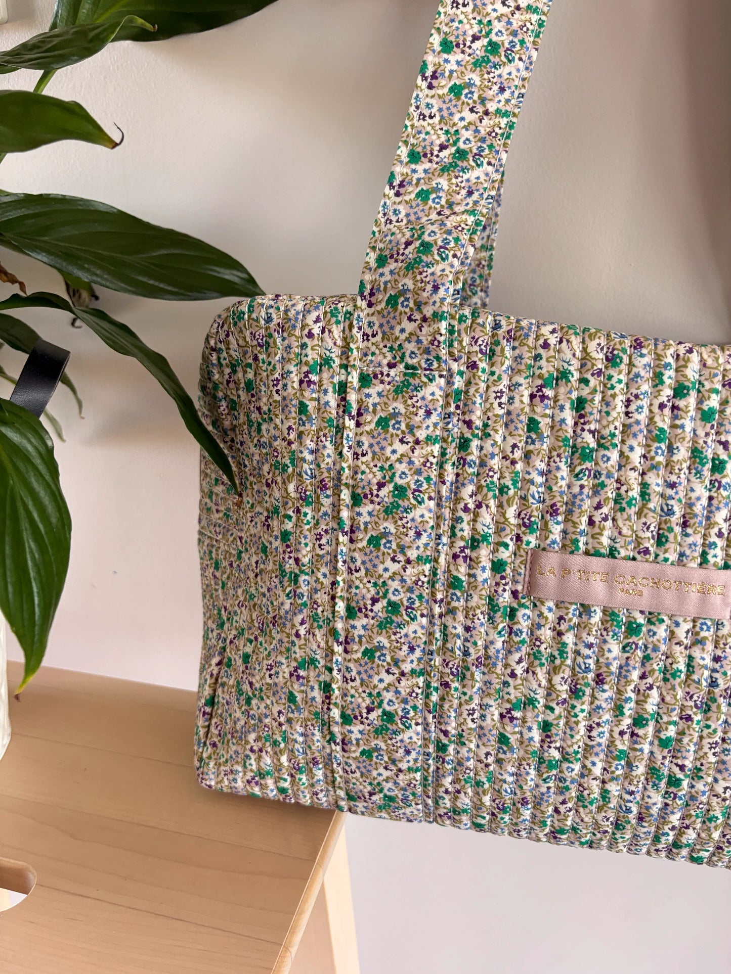Sac polochon vert à motif floral, matériau matelassé, avec bandoulière et petit label rose sur le côté.
