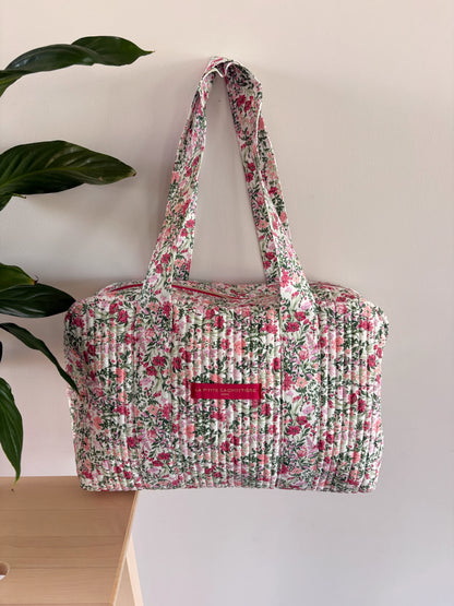 Sac polochon matelassé floral rose et vert sauge sur fond blanc avec poignées longilignes et étiquette rose.