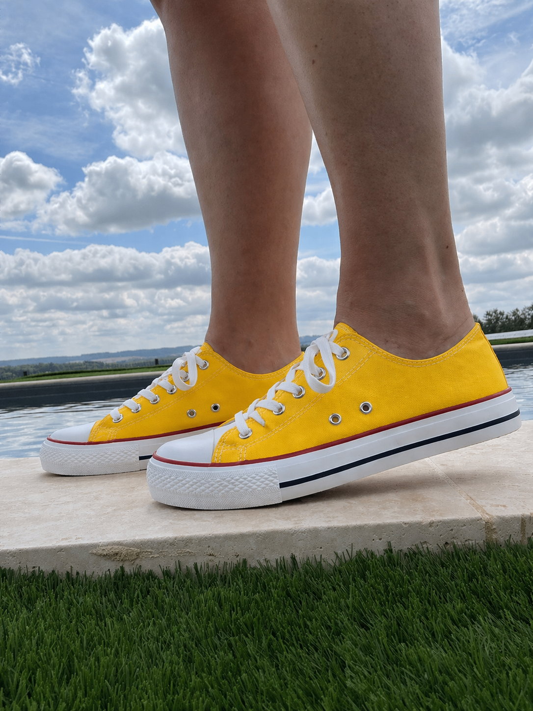 Paire de baskets jaunes Converse style basse, lacets blancs, vue pieds en extérieur sur une dalle près de l’eau.