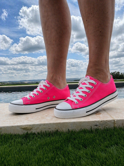 Baskets Converse roses fluo à brides blanches, pieds posés sur un rebord près d’un lac par temps nuageux.