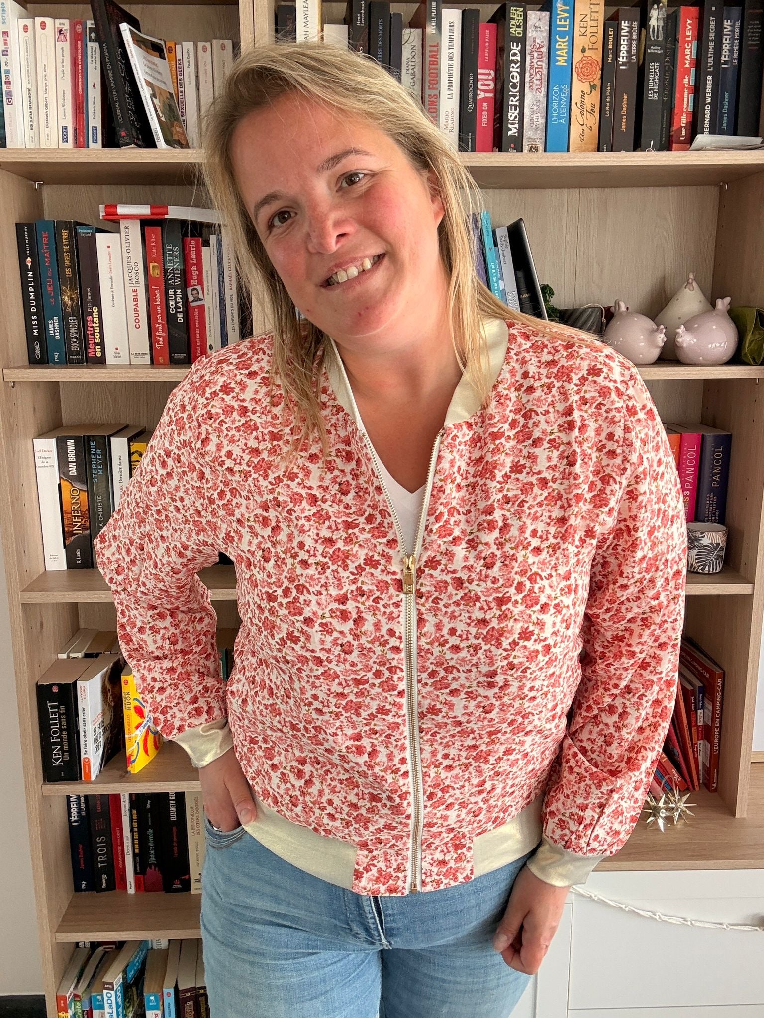 Veste bomber légère à fleurs rouges et orangées portée par une femme, fond bibliothèque, pose décontractée.