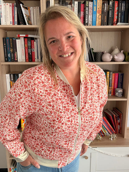 Veste légère fleurs rouges et rosées, zippée, portée par une femme souriante dans un intérieur avec bibliothèque.