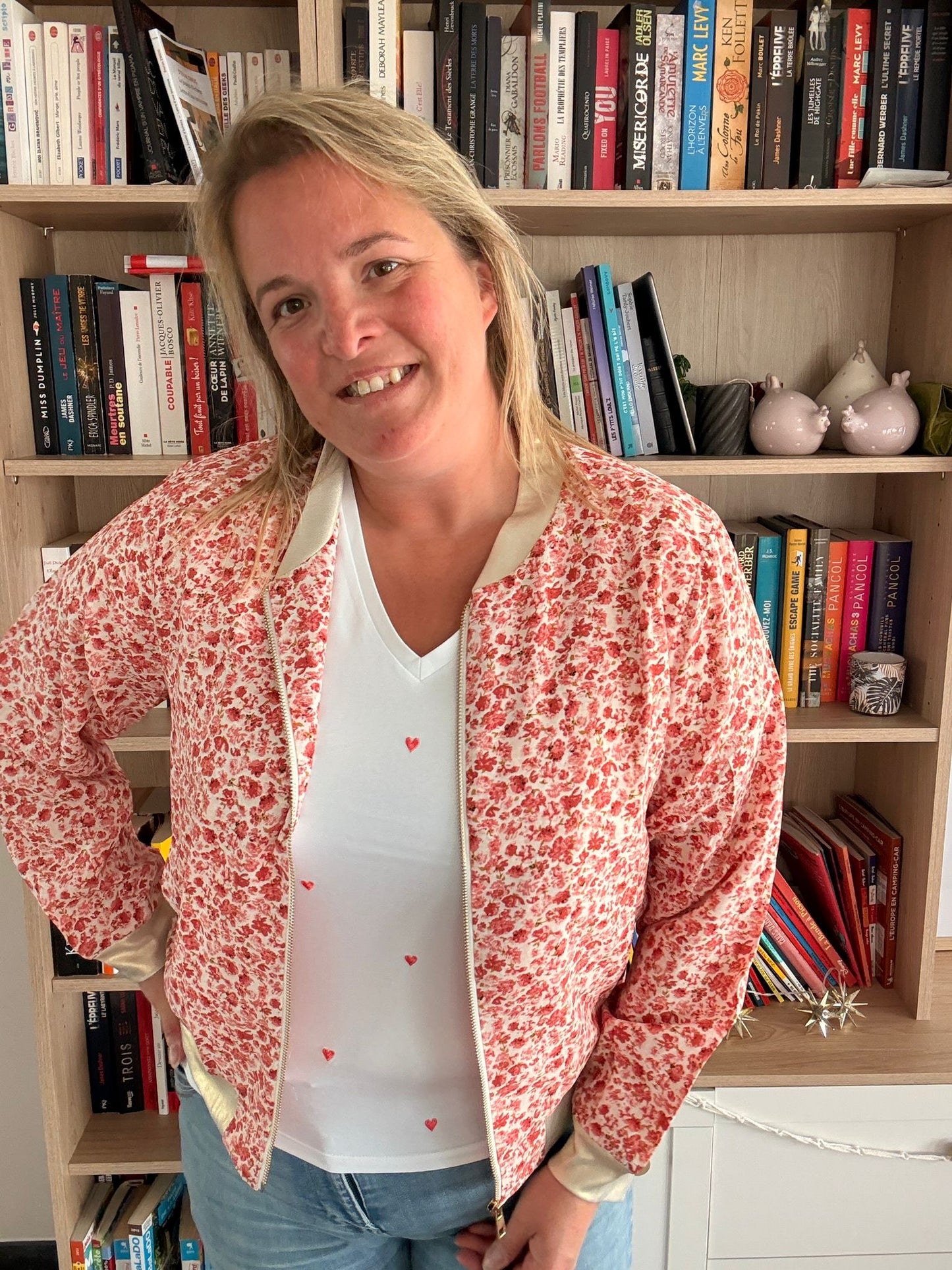 Veste bomber légère à motif fleuri rouge et orangé portée par une femme souriante devant une bibliothèque.