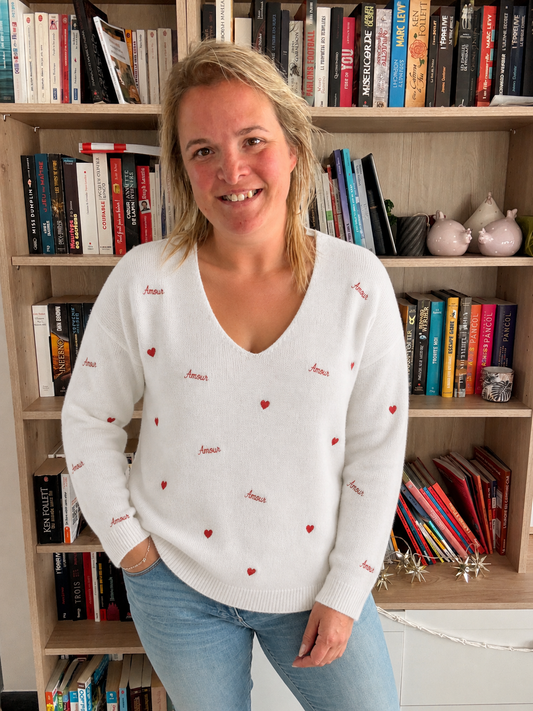 Pull amour brodé blanc porté par une femme souriante dans une bibliothèque, pull-over blanc orné de petits cœurs et du mot « Amour ».
