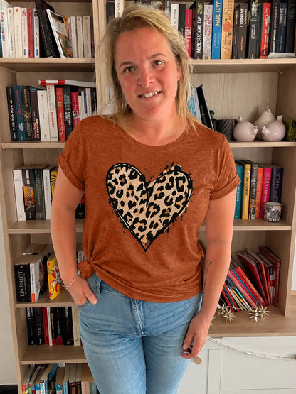 Homme ou femme portant un t‑shirt terracotta à cœur léopard, debout dans une bibliothèque, les mains dans les poches.