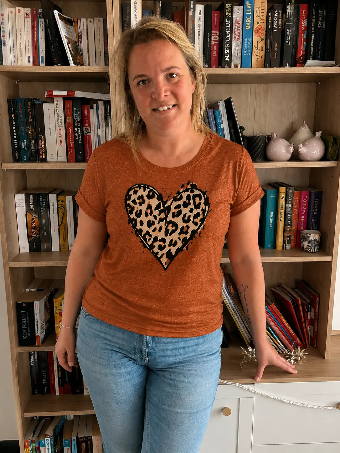 Homme/femme portant un T-shirt terracotta imprimé cœur léopard, debout près d’une bibliothèque.