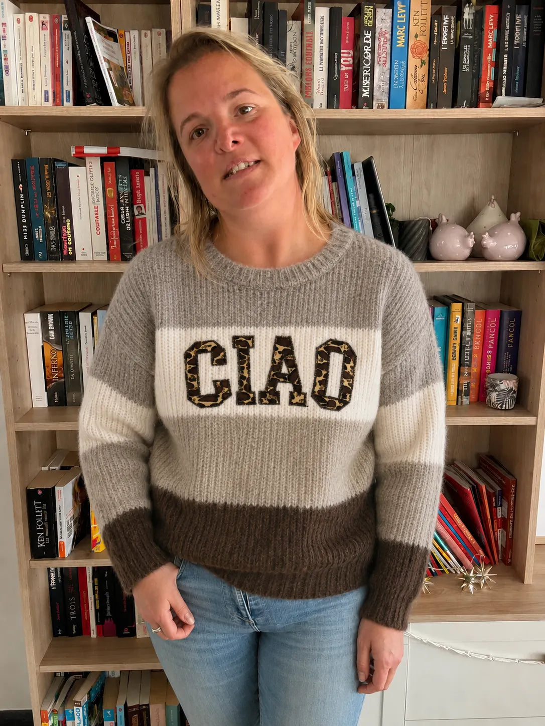 Pull en maille femme CIAO gris marron portée par une femme devant une étagère remplie de livres.