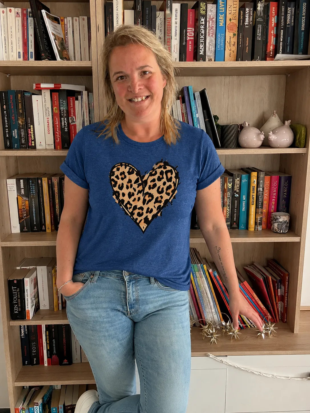 T-shirt bleu femme avec cœur léopard imprimé graphique chic, porté par une femme souriante debout devant une bibliothèque.