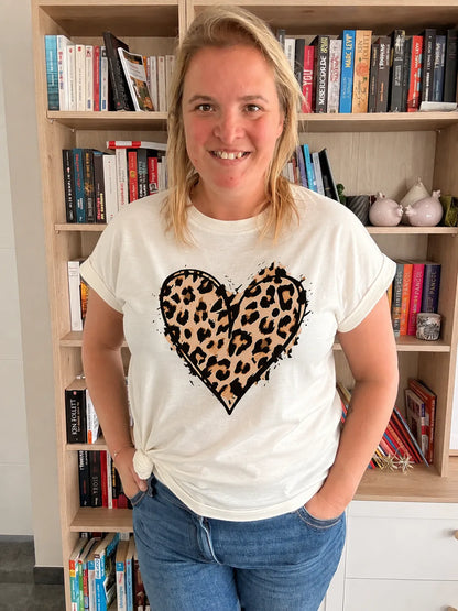 T-shirt blanc femme avec cœur leopard imprimé sur le torse, col rond, manches courtes, portée par une femme souriante dans une pièce avec bibliothèque en arrière-plan.