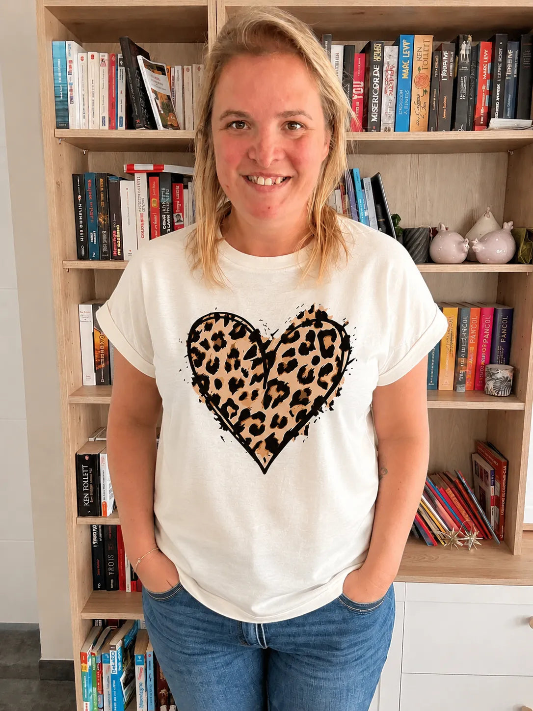 T-shirt blanc femme avec cœur imprimé léopard, col rond et manches courtes, portée par une femme devant une étagère de livres.